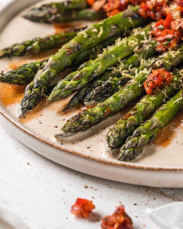 close up of asparagus with Turkish Ezme inspired Tomato Salad and Creamy Tahini Dressing on a white plate with a white wood background
