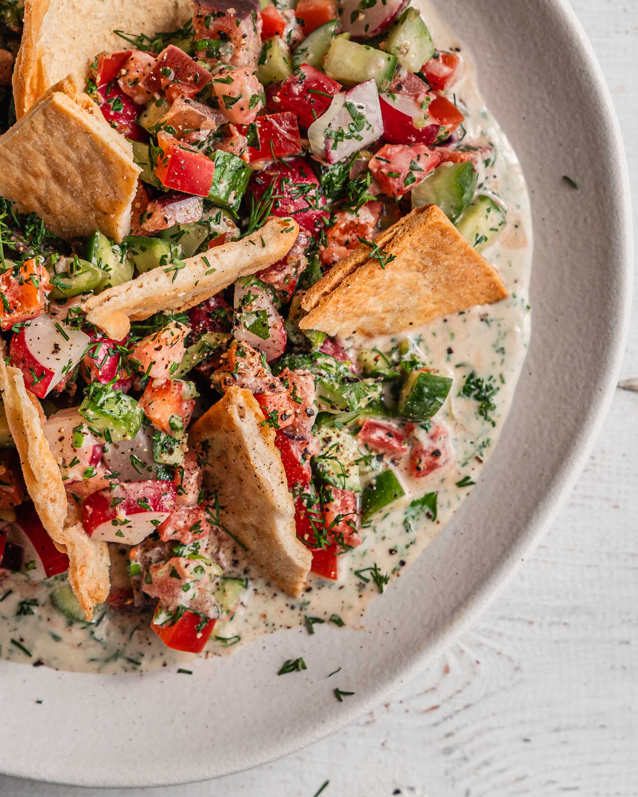 A white plate overflowing with a colorful chopped salad featuring chopped vegetables, fresh herbs, and a dollop of creamy tahini dressing. Crispy pita chips are scattered throughout the salad for added texture.